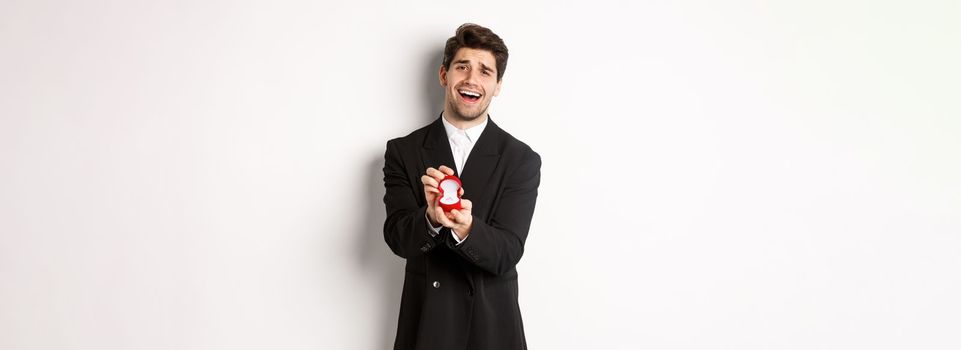 Portrait of handsome man in black suit, open box with wedding ring, making a proposal, asking to marry him, standing against white background.