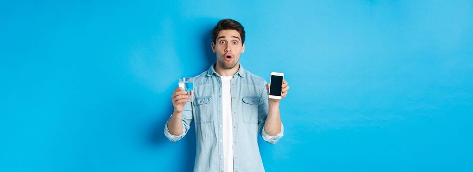 Man looking surprised, showing smartphone screen and glass of water, standing over blue background.