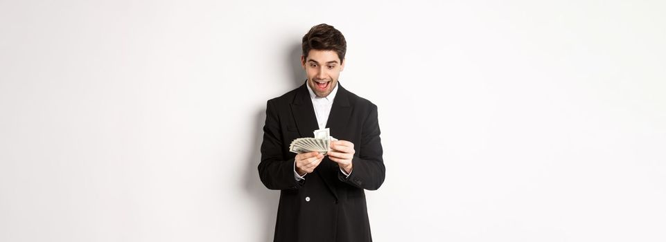 Image of excited handsome businessman, counting money and smiling amused, standing against white background in suit.