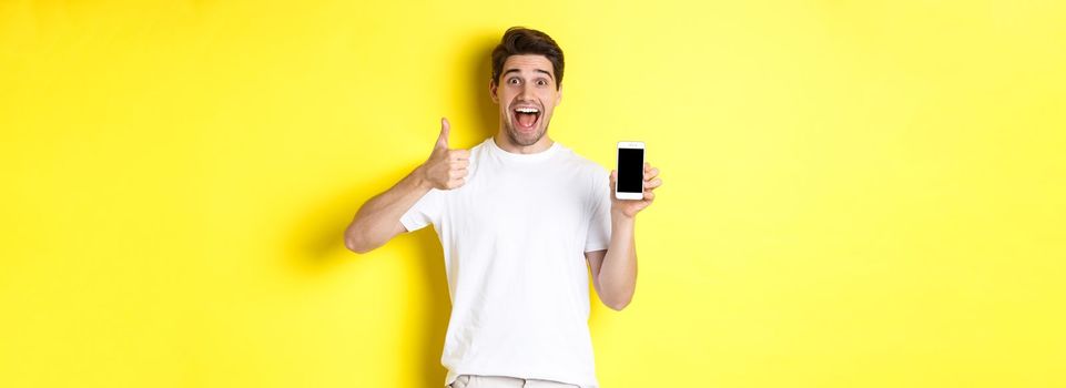 Happy young man showing thumb up and mobile phone screen, recommending application or internet website, standing over yellow background. Copy space