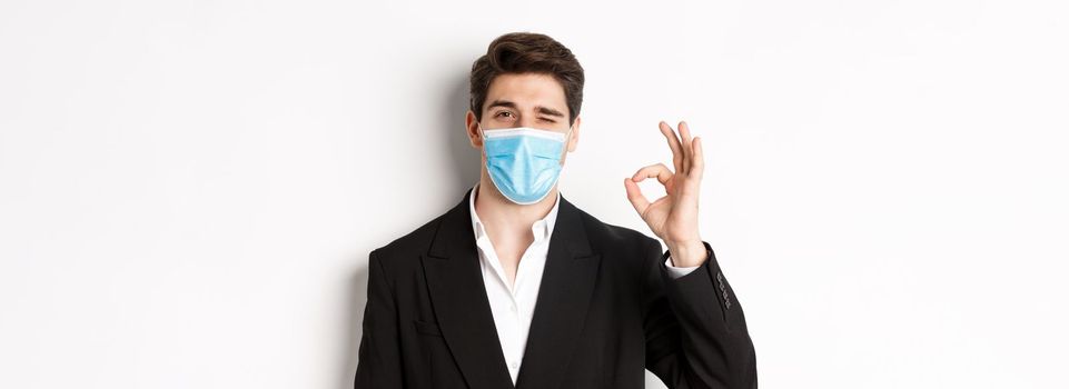 Concept of covid-19, business and social distancing. Close-up of good-looking businessman in trendy suit and medical mask, showing okay sign and winking, standing against white background.