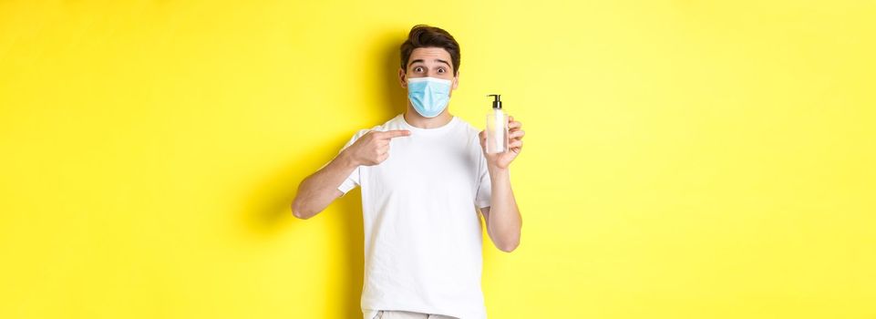 Concept of covid-19, quarantine and lifestyle. Excited guy in medical mask showing good hand sanitizer, pointing finger at antiseptic, standing over yellow background.