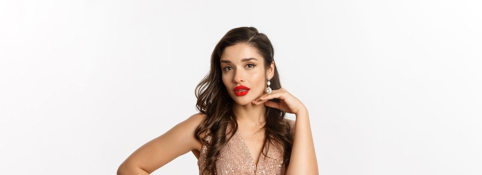 Concept of New Year celebration and winter holidays. Close-up of beautiful elegant woman with red lips and party dress, touching her face and looking at camera, white background.