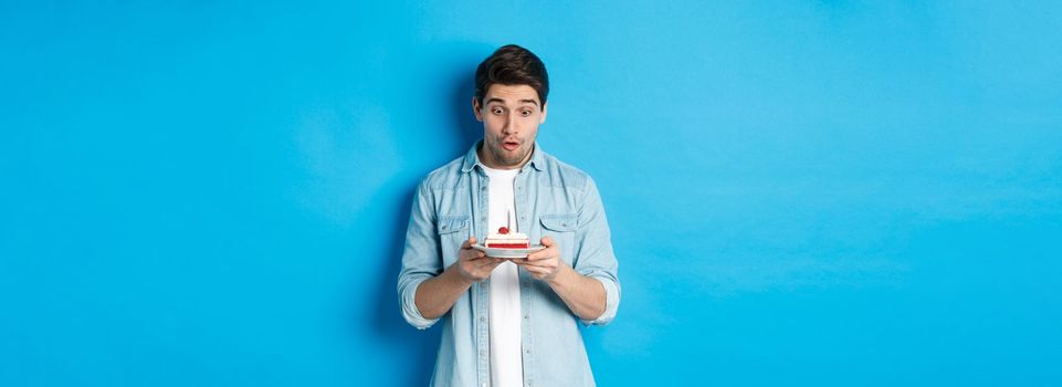 Birthday. Surprised man looking at b-day cake, standing against blue background.