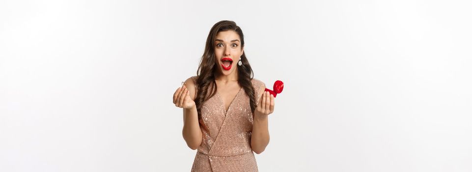 Excited woman holding engagement ring and red box, looking surprised and amazed, receive marriage proposal on date, wearing elegant dress, standing over white background.