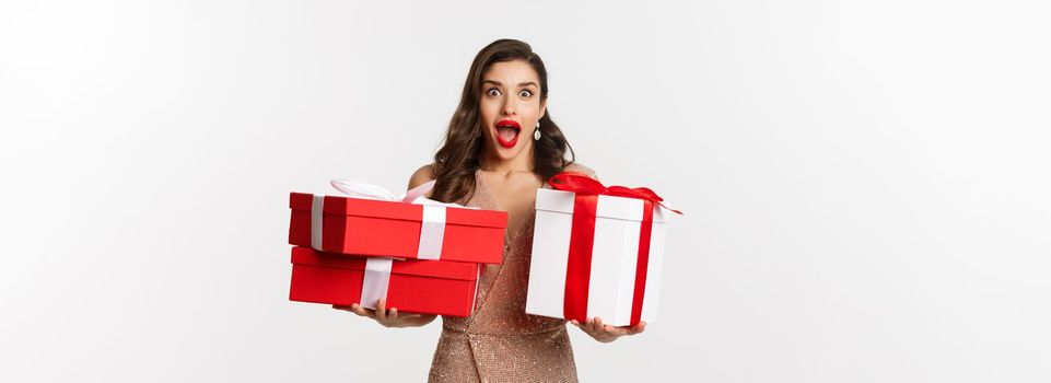 Holidays, celebration concept. Excited and surprised woman holding Christmas gifts and smiling amazed, wearing glamour dress, standing over white background.