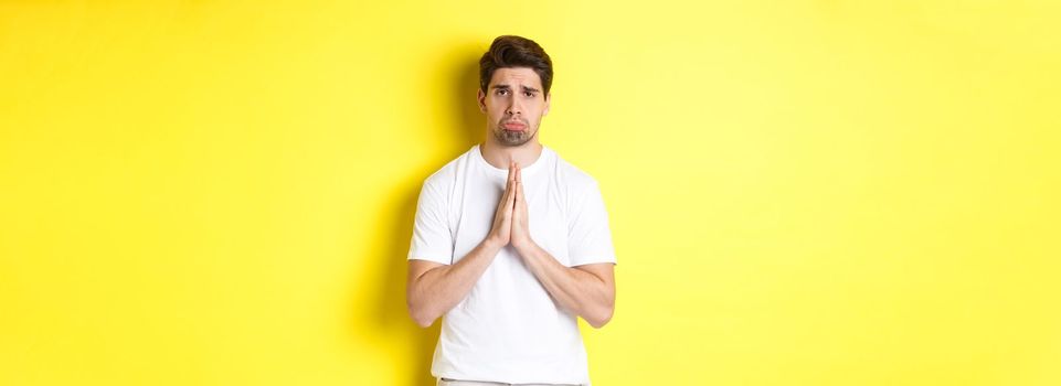 Sad guy begging for something, sulking and asking for favour, saying sorry, standing over yellow background.