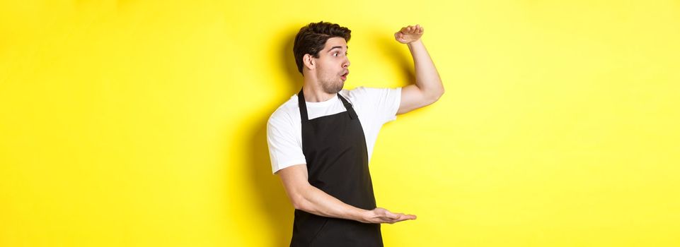 Waiter in black apron looking amazed at something large, holding big object, standing over yellow background.