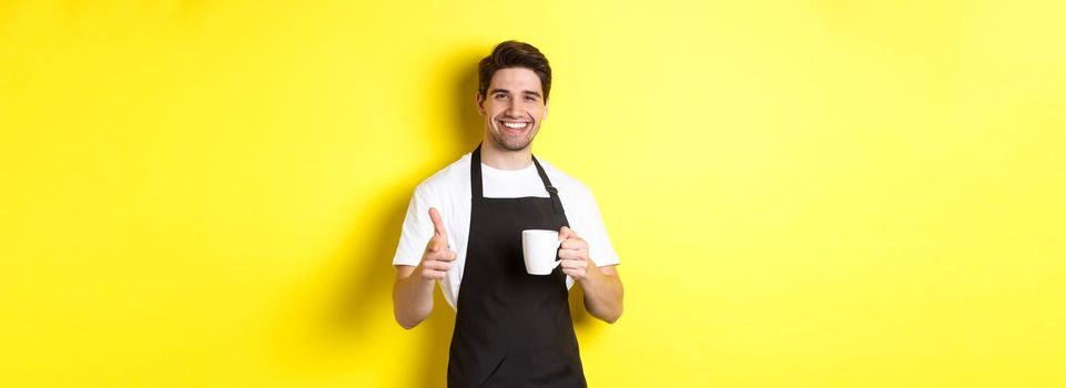 Barista bringing coffee and pointing finger gun at camera, standing in black apron against yellow background.