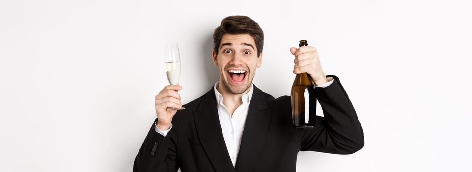 Close-up of handsome smiling man in black suit, making a toast, holding champagne and glass, celebrating new year, standing against white background.