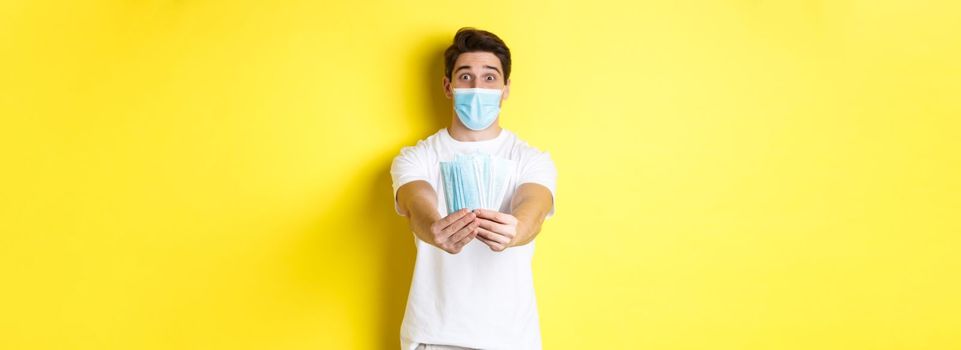 Concept of covid-19, quarantine and preventive measures. Young caucasian man giving medical masks for you, standing against yellow background.