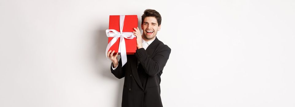 Concept of christmas holidays, celebration and lifestyle. Attractive man in black suit, holding new year gift and smiling, standing with a present over white background.