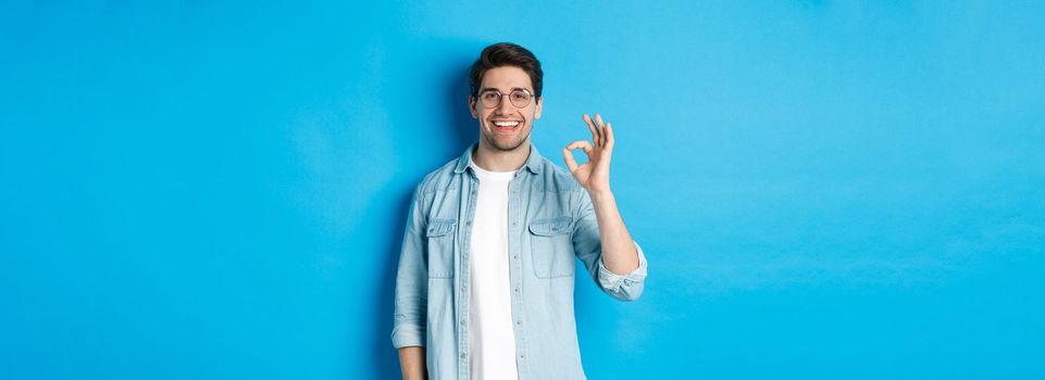 Attractive young man wearing glasses and casual clothes, showing ok good sign in approval, like something, standing against blue background.