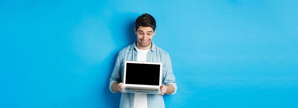 Excited handsome man looking at screen, showing promo offer on computer screen, smiling amazed, standing over blue background.