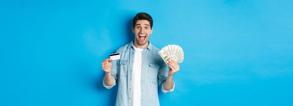 Happy attractive man looking amazed, showing cash and credit card, concept of banks, credit and finance. Blue studio background.