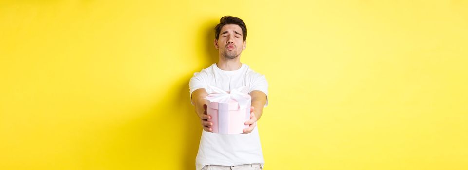 Concept of holidays and celebration. Boyfriend giving a gift, close eyes and pucker lips for kiss, standing over yellow background.