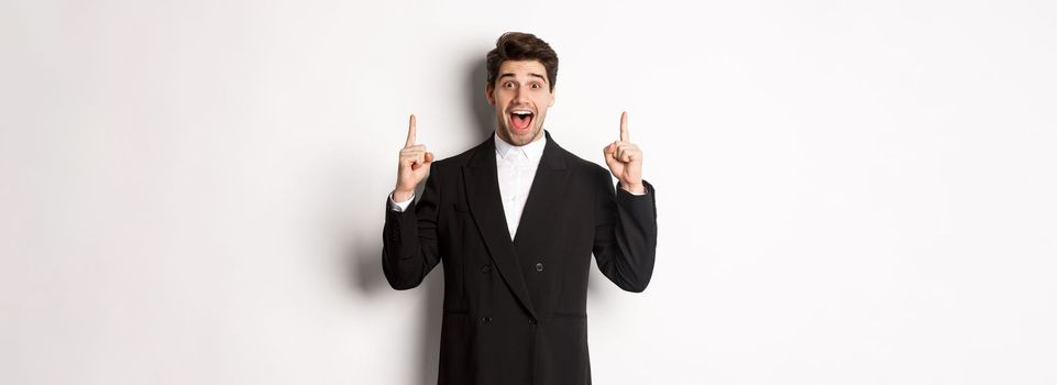 Image of handsome man in party suit, showing holidays promo, pointing fingers up and smiling amazed, standing over white background.