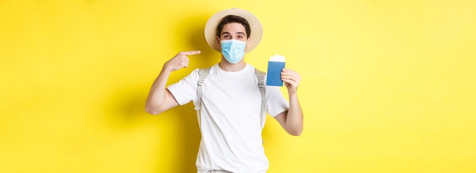 Concept of covid-19, tourism and pandemic. Man tourist showing passport, travelling in medical mask for protection from coronavirus, yellow background.