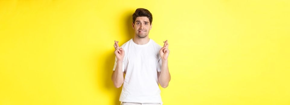 Nervous guy crossing fingers for good luck, hoping for something, standing over yellow background.