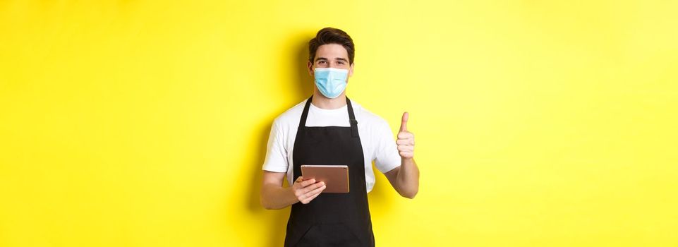 Concept of covid-19, small business and pandemic. Friendly waiter in medical mask and black apron showing thumb up, taking orders with digital tablet, yellow background.