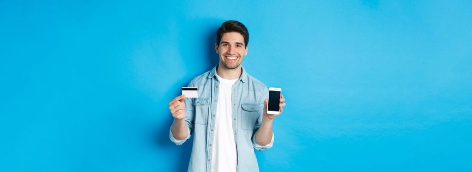 Young smiling man showing smartphone screen and credit card, concept of online shopping or banking.