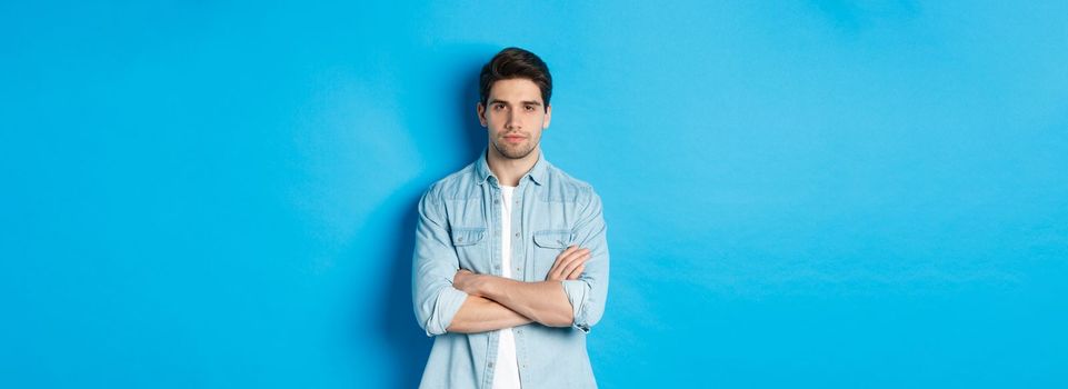 Image of handsome caucasian man in casual outfit, looking serious and confident, standing against blue background.