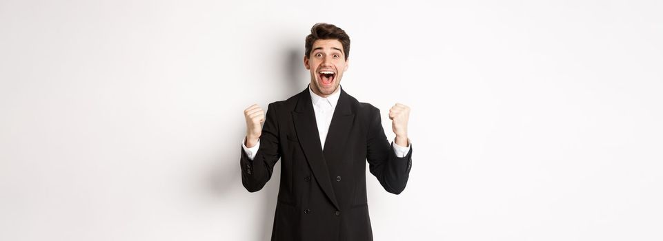 Image of happy and relieved businessman feeling lucky, making fist pumps and smiling with joy, achieve goal, winning and triumphing, standing over white background.
