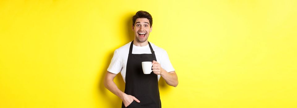 Smiling young barista in black apron holding coffee cup, standing over yellow background.