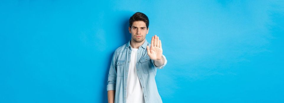 Serious man frowning and saying no, extending hand to shop stop sign, prohibit action, standing against blue background.