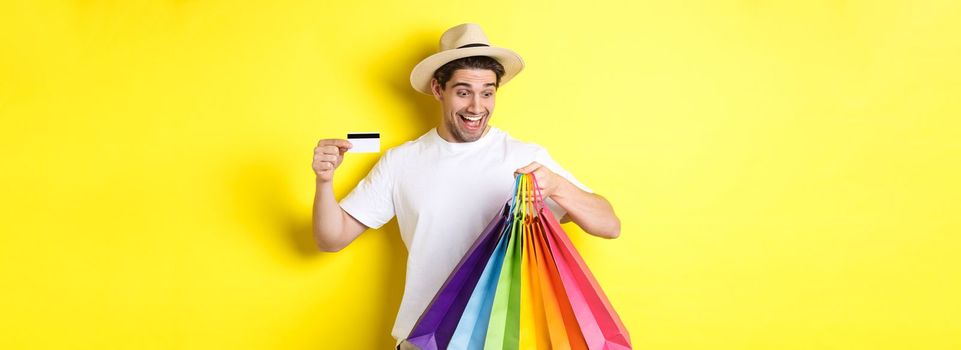 Concept of vacation and finance. Happy man shopper looking at shopping bags satisfied, showing credit card, standing against yellow background.