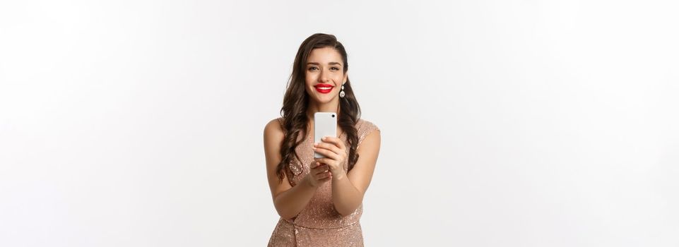 Beautiful young woman in party dress and makeup, taking photo on smartphone, shooting you on phone camera, standing over white background.