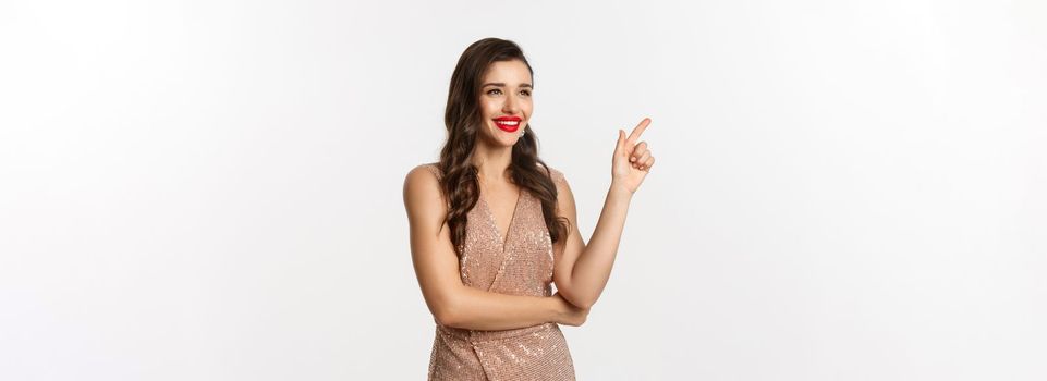 Christmas, holidays and celebration concept. Attractive woman wearing party dress and red lipstick, pointing and looking left, enjoing new year eve, white background.