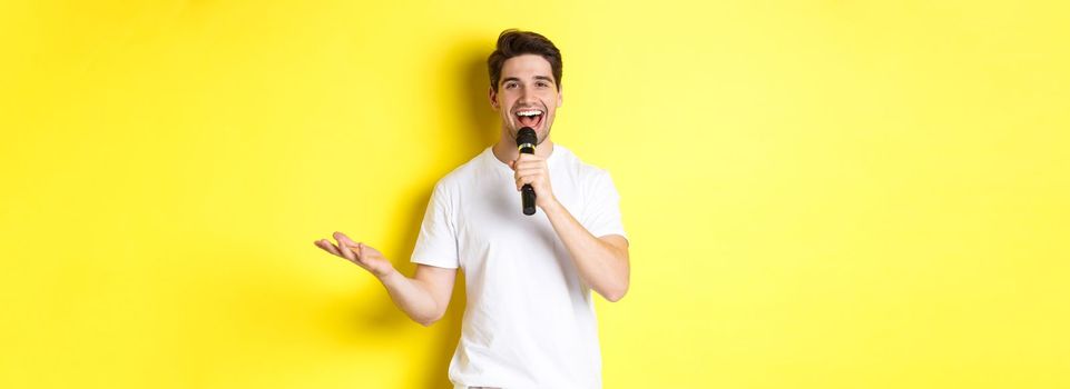 Happy man entertainer performing, talking in microphone, making speech or stand-up show, standing over yellow background.