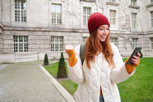 Cheerful redhead girl winning, celebrating victory, read great news on mobile phone and jumping from joy, saying yes, standing outside building in yard.