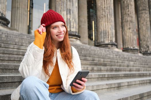 Stylish young redhead woman, talking on mobile phone app, using social media application, looking for something online on smartphone, sits on stairs outdoors.