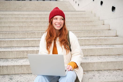 Stylish young redhead girl, student with laptop sitting on stairs near building, connects to public wifi and works on project, chats on computer.