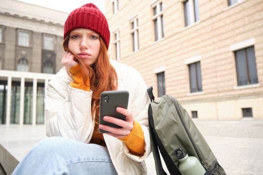 Redhead 20s years girl, sits with smartphone outside building, looks complicated and upset, frowns thoughtful, holds mobile phone in hand.