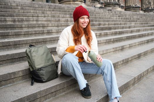 Smiling tourist, girl sits on stairs, rests on staircase, takes thermos from backpack, drinks hot coffee from flask.