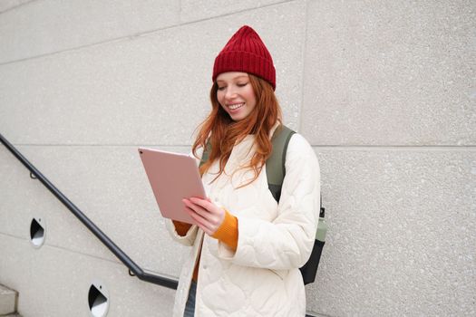 Young redhead woman with red hat, uses her digital tablet outdoors, stands on street with gadget, connects to wifi internet and searches for a location in internet.