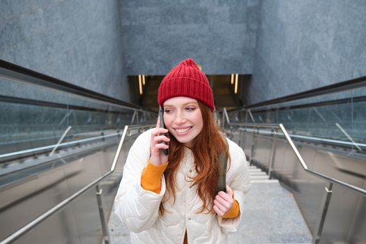 Portrait of happy redhead woman walking around town with smartphone, calling someone, talking on mobile phone outdoors.