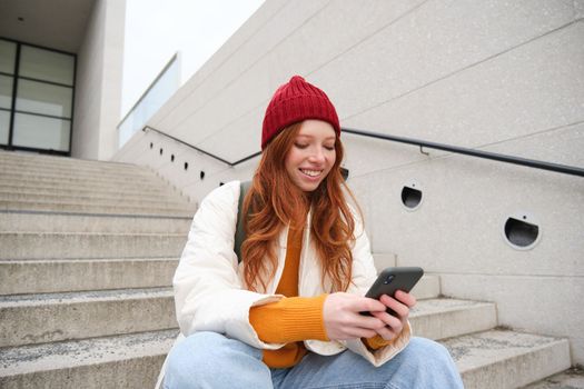 Redhead girl in red hat, sits on stairs and uses mobile phone. Modern woman holding smartphone, texting message, using telephone application outdoors.