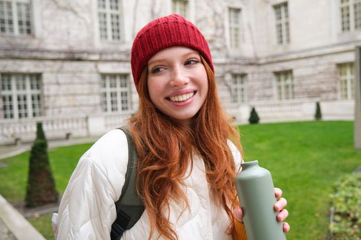 Portrait of happy young woman, tourist with backpack sightseeing, drinking hot tea from thermos, holding flask and smiling. Tourism and people concept