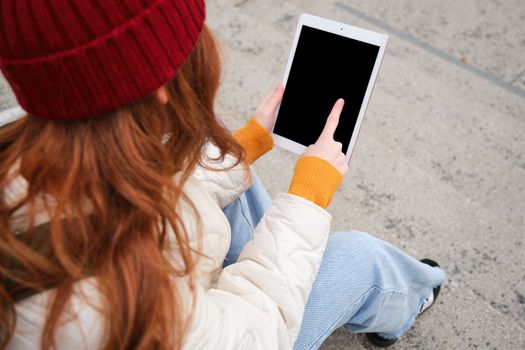 Rear view of redhead girl touches digital tablet screen, touchpad, texts message, uses internet application on gadget, sits on stairs outdoors.