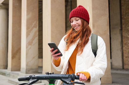 Redhead girl, tourist with backpack, uses mobile phone to rent e-scooter on streets of European city.