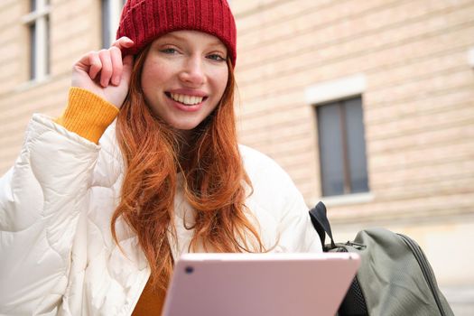 Beautiful redhead woman in red hat, sits with backpack and thermos, using digital tablet outdoors, connects to wifi, texts message, books tickets online.