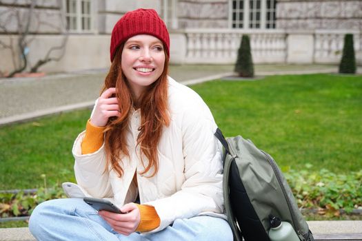 Redhead girl, female student sits with mobile phone on bench in parj, leans on her backpack. Woman browsing social media app feed on her smartphone.