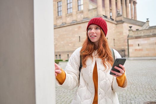 Smiling redhead girl tourist, walks around city and explores popular landmarks, sightseeing, holding smartphone, looking at her mobile app and checks with city map.