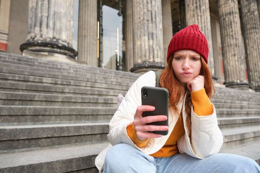 Portrait of young redhead woman with complicated face, sits on street stairs in red hat, holds smartphone and frowns thoughtful, feels uneasy.