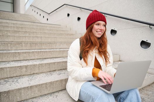 Smiling redhead girl, student sits on stairs outdoors and uses laptop, connects to public wifi in city and works on project, uses internet on computer.