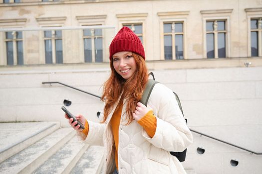 Joyful girl tourist looks at mobile phone, texts message on smartphone social media application, walks around city, looks for sightseeing on mobile app.